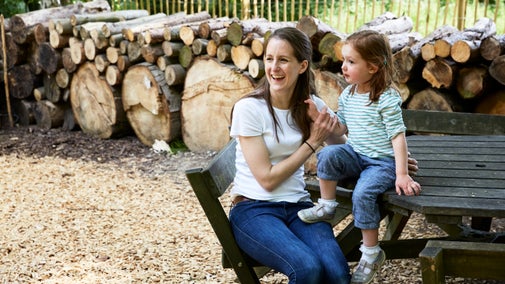 An adult and child sit at a picnic table in a children's play area with piles of logs behind them at Anglesey Abbey, Cambridgeshire